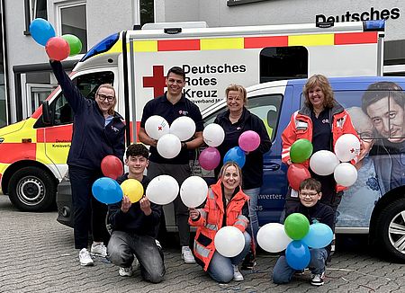 Auf dem Bild sind fröhliche DRK'ler mit einem Luftballon zu sehn. Im Hintergrund seiht man ein Rotkreuzfahrzeug. R