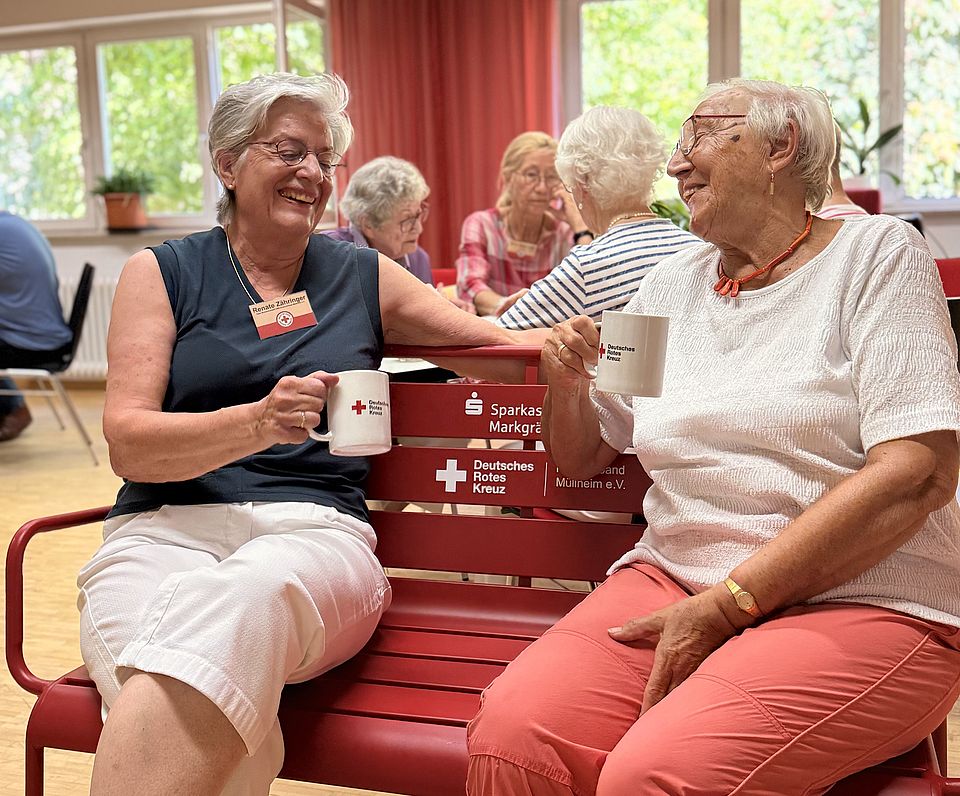 Auf dem Bild sind zwei Seniorinnen auf einer roten Bank zu sehen.
