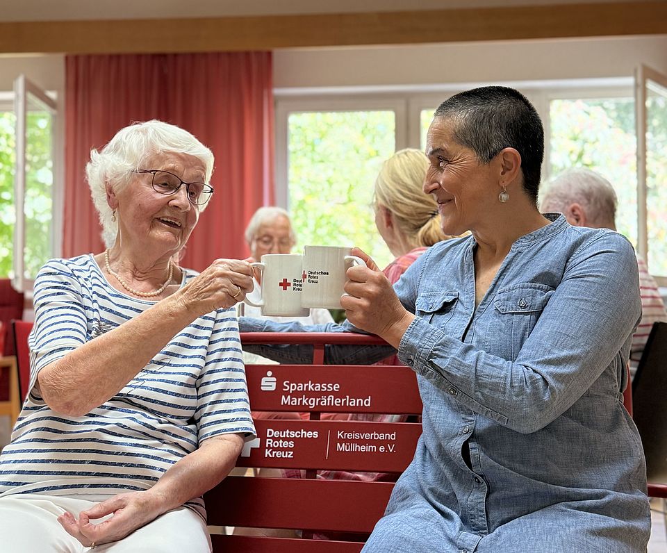 Auf dem Bild sind zwei Seniorinnen auf einer roten Bank zu sehen.