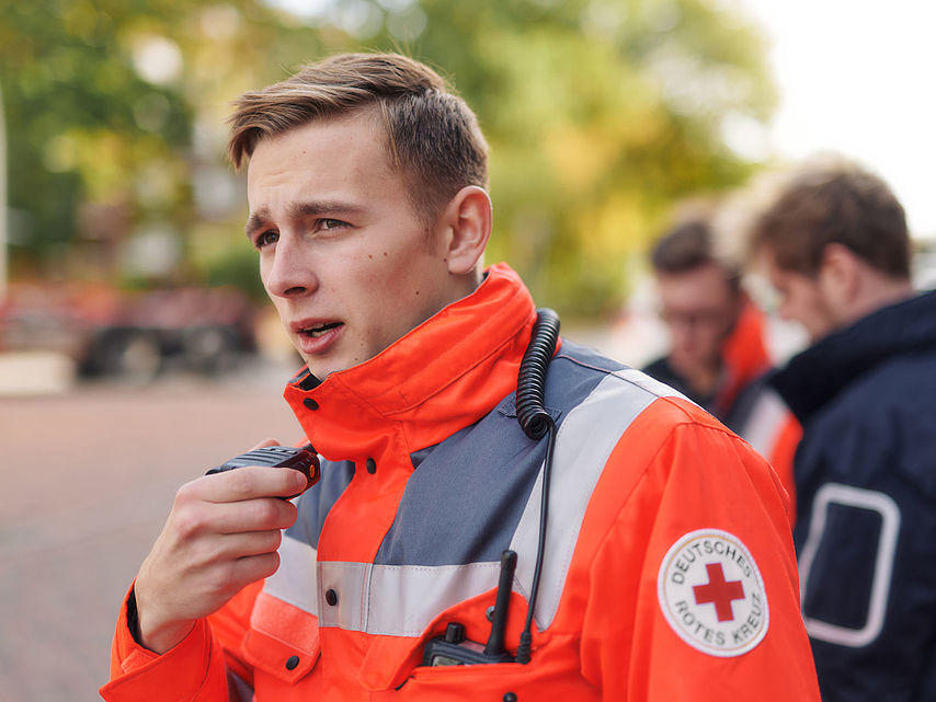 Symbolfoto, Mitarbeiter des DRK in Einsatzjacke spricht in ein Funkgerät
