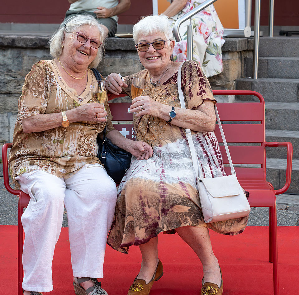 Rote Bank der Begegnung_Kino&Wein Auf dem Bild sind zwei Damen auf einer roten Bank zu sehen.