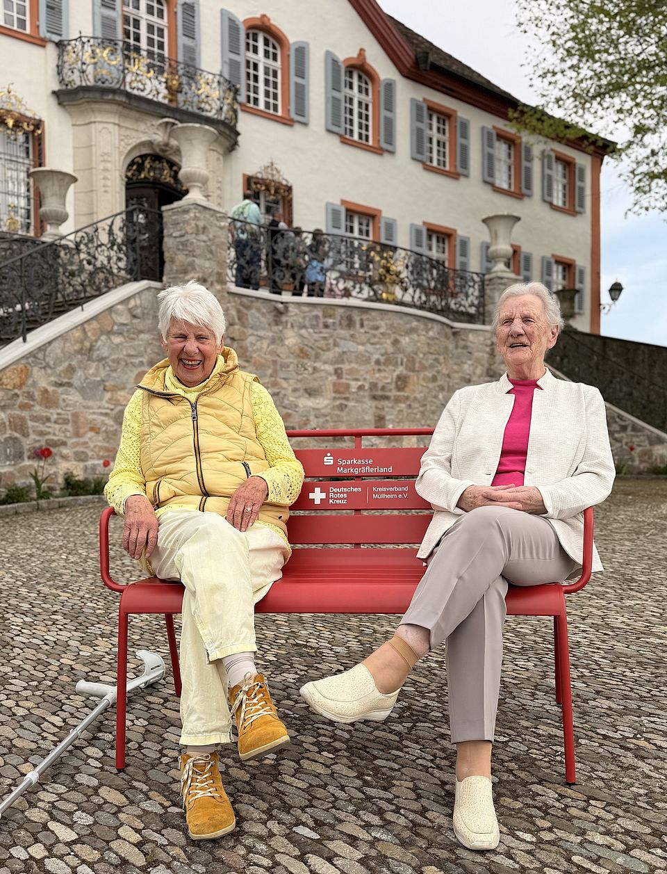 Rote Bank der Begegnung-Schloß Bürgeln Auf dem Bild sind 2 Damen auf einer roten Bank zu sehen.
