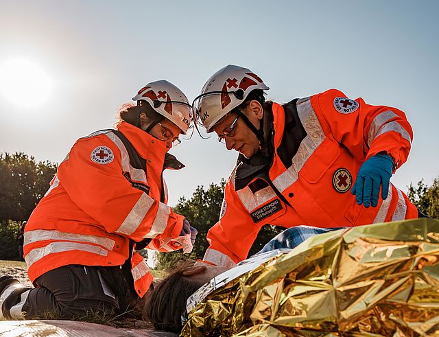 Zwei ehrenamtliche aus einer Bereitschaft helfen einer verletzten Person und breiten eine Rettungsdecke über sie aus.